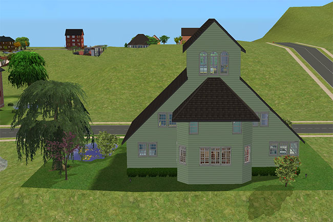 A rear view of Moonlit Bay shows pale green siding, layered roof sections, tall upper windows, a bay extension, side landscaping, a nearby pond, and neighborhood roads beyond the lot.