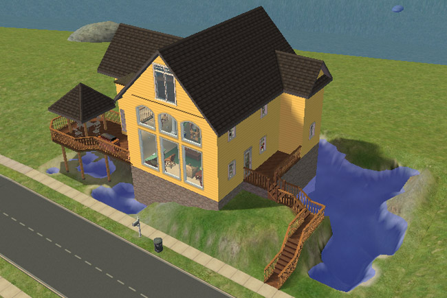 An angled view shows the yellow house beside the road, with decks, stairs, rocky landscaping, and blue water along the side and back.